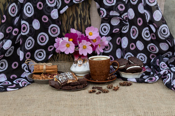 On the table is a bouquet of cosmos, a cup of coffee with lemon, on saucers is a piece of cake and chocolate, cookies, cinnamon sticks, almonds, peanuts and coffee beans..
