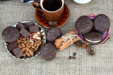 There is a cup of coffee on the table, on the saucers are chocolate, cookies, cinnamon sticks, almonds, peanuts, star anise and coffee beans..
