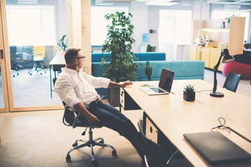 Experienced businessman is sitting near his workplace in the office while thinking is looking away. Mature male entrepreneur deep in thought is taking break after hard work on portable laptop computer