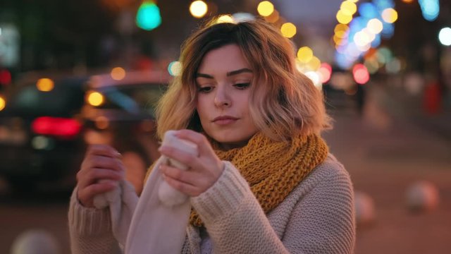 Portrait frozen young woman knitted scarf rubs her hands and puts on warm knitted mittens of bokeh of Christmas lights and city street scenery on walk in evening in winter. Noel. Emotions. Lifestyle