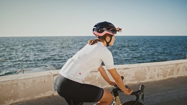 Sportswoman in protective helmet and sportswear is riding trekking bike along sea embankment. Sunny summer day, blue sky and sea. Slow motion