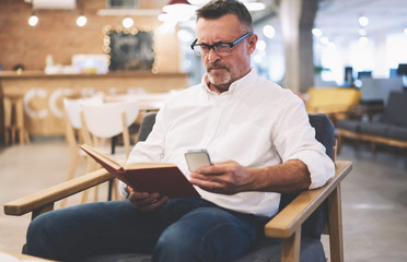 Handsome male dressed in trendy clothes fond of reading enjoying leisure spending with book taking photos of favorite paragraph to share with followers in social networks using phone connected to wifi