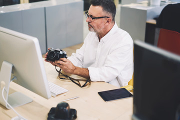 Professional male employee in eyeglasses sitting at computer and making photos for productive work.Mature man in optical spectacles taking pictures on stylish vintage camera in coworking space
