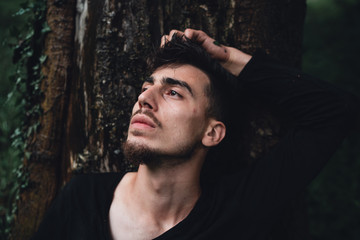 Young man leaning near a tree in the forest thinks