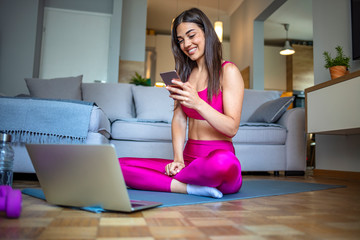 A sporty woman in sportswear is sitting on the floor with dumbbells and a bottle of water and is using a laptop and smart phone at home in the living room. Sport and recreation concept.