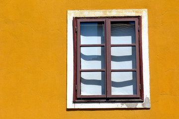 Fototapeta na wymiar painted brown wooden window in vertical orientation in yellow color stucco exterior wall. bright summer light. European traditional facade detail in old town. residential home. shadows and lights. 