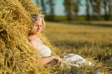 A woman in a white dress in a haystack at sunset. An evening of peace and quiet. A mother of three boys, a wonderful figure. Light hair, light clothing, spikelets in the field. Belarus.