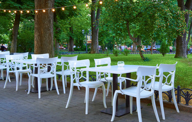 open air street cafe outdoor - white chairs and tables, decorated with illumination, the city Park and walking people