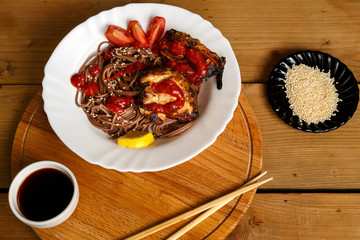 Soba with chicken and vegetables in a white plate on a stand with soy sauce and sesame seeds next to sticks.