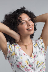 beautiful young brunette woman with curly hair posing with raised arms, wearing jewelry and a white patterned shirt