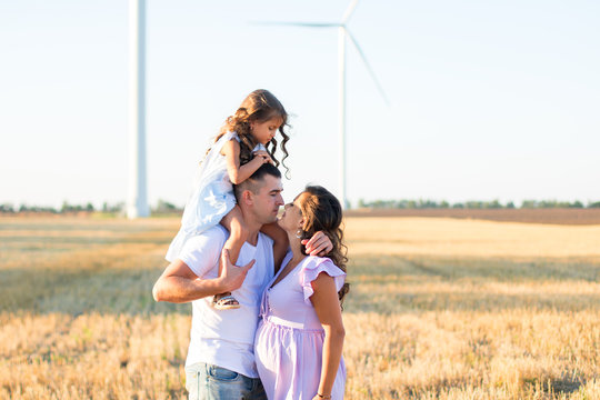 young husband kissing his pregnant wife and a little girl on the father's shoulders on the field, wind turbines on the background