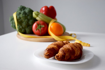 Plate with fresh baked croissants and set of vegetables with tape measure on background. Choosing between bad food and healthy eating. Balanced diet, clean eating or weight loss concept.
