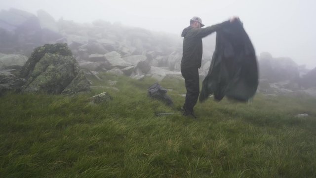 A man setting up a camping tent on a misty windy mountain