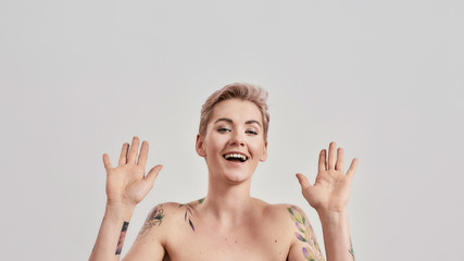 Portrait of half naked tattooed woman with short hair laughing at camera, raising her hands isolated over light background