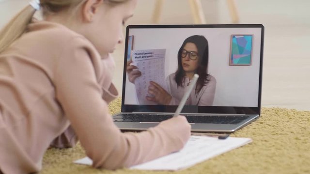 Back view of Caucasian schoolgirl with blond hair is lying on carpet in living room, having video meeting with young female teacher and doing math task