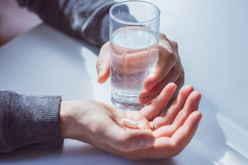 Image of a young man holding a glass of water in one hand and a white tablet in the other. The man is dressed in a gray sweater