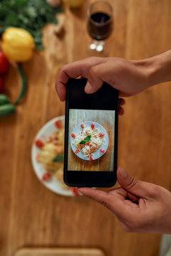 Top view of hands of man, cook taking picture of garlic butter shrimp pasta, meal decorated with basil and cherry tomatoes. Cooking at home, Mediterranean cuisine concept