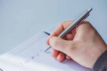 An image of a man's hand writing the phrase "I can do it!"in a Notepad with a ballpoint pen. The man is sitting at a white table and is dressed in a gray jacket.