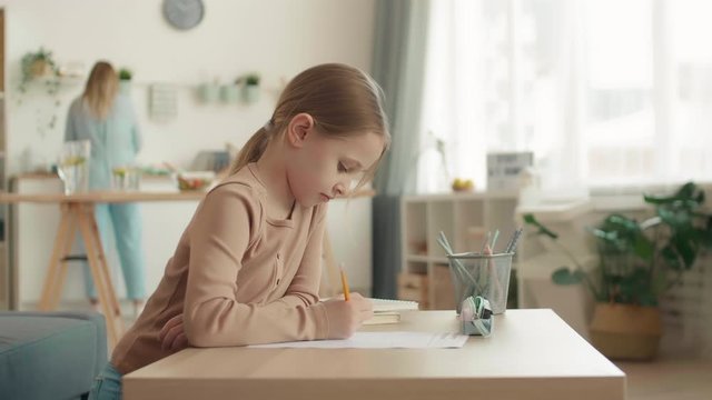 Lockdown portrait of Caucasian schoolgirl with blond hair is sitting at desktop at home, doing difficult task and then looking at camera while her mom cooking on background