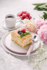 Slice cake of puff pastry with mint, raspberry and blueberry on plate, cup of coffee, peony. Delicate and airy composition, vertical format.