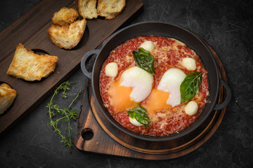 hearty English breakfast - poached eggs, with tomatoes and sauce in a pan on a wooden board
