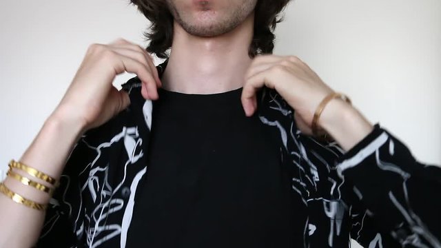 Young caucasian man putting on black and white pattern shirt, wearing gold color bracelets, black t-shirt underneath