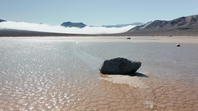 Close up of black rock on the wet cracked surface of Death Valley desert. Epic race track playa park. Cinematic 4K aerial of moving stones on sunny morning, with mountains and clouds on background