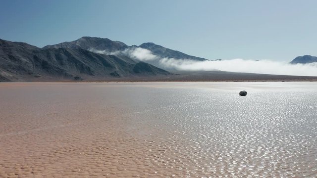 Drone flying around black rock on wet cracked surface of Death Valley desert. Epic race track playa park. Cinematic 4K aerial of moving stones on sunny morning, with mountains and clouds on background