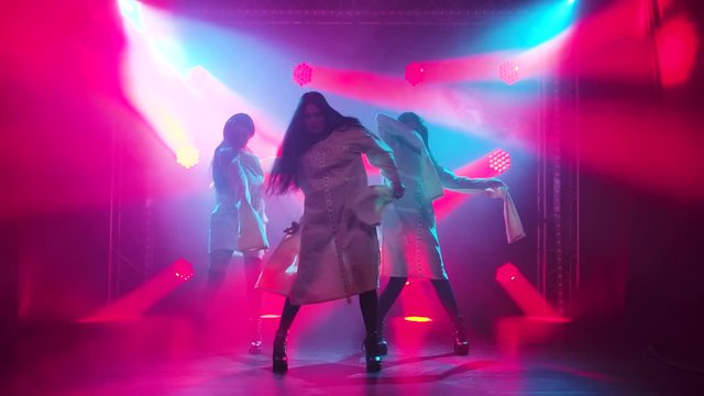 Three charming girls in white straitjacket are dancing on the stage. Silhouettes in a dark studio with smoke and neon lighting. Red neon light effects. Dance show. Slow motion.