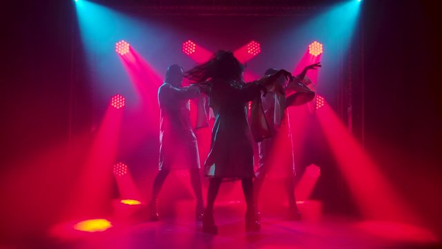 Three charming girls in white straitjacket are dancing on the stage. Silhouettes in a dark studio with smoke and neon lighting. Red neon light effects. Dance show. Slow motion.