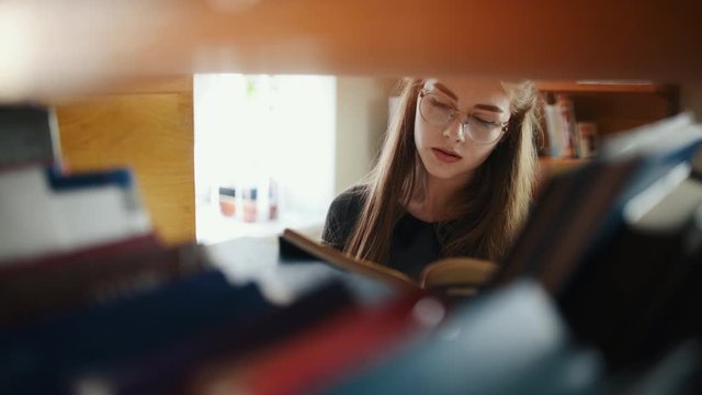 View through the shelves of female student in eyewear that searching books in library.