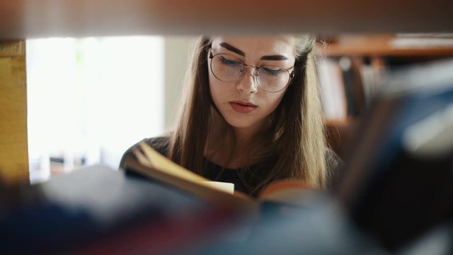 View through the shelves of female student in eyewear that searching books in library.