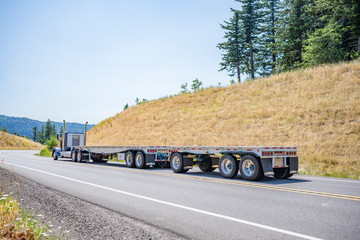 Blue big rig day cab semi truck tractor transporting two empty flat bed semi trailers driving on the winding road at Columbia Gorge