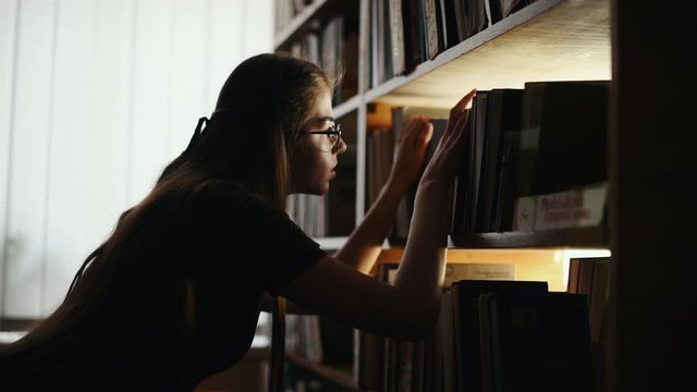 Girl in eyewear searching for the book and looks at the light beam through the shelves.