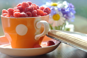 full ceramic colored teaCup of raspberries on saucer and corner of open book on blurry background of bunch of flowers. Beautiful summer berries wallpaper