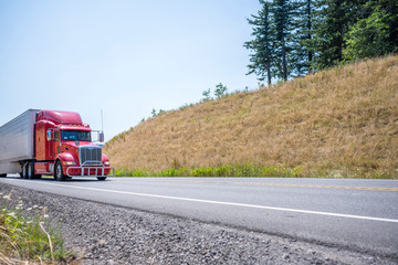 Fototapeta na wymiar Powerful red big rig diesel semi truck carry cargo in refrigerator semi trailer running on the summer winding road with hill and trees