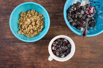 Blue bowls of crumble topping and blackberries in sugar/flour mix, white bowl, on a wood background
