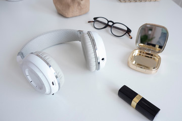 white wireless headphones, glasses, lipstick and a mirror on a white table..