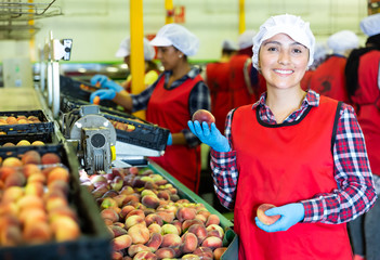Smiling Latina woman worker standing in fruits industrial production facility holding ripe peaches