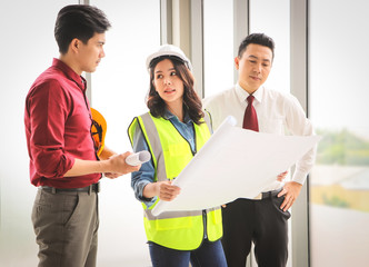  three construction workers, one woman and two men standing by windows , holding blue print and  discussing about  project.