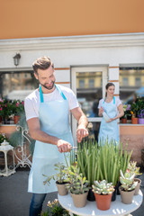 Bearded male cutting leaves and brown-haired female standing with water spray outside flower shop