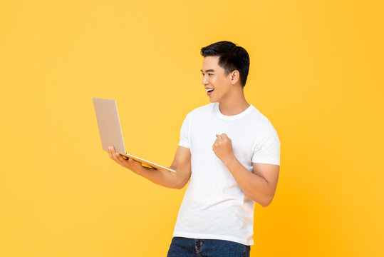Portrait of a smiling  young handsome Asian man holding laptop while doing a winning closed fist gesture in isolated studio yellow background