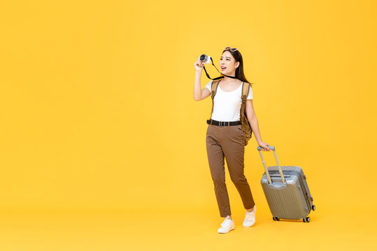 Full length travel portrait of excited young tourist Asian woman walking while taking photo with camera in isolated studio yellow background