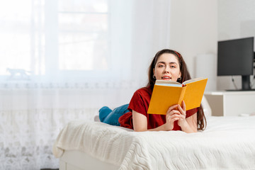 A young Caucasian woman is lying on the bed on her stomach and holding a book with a smile. Copy space. White interior of the room on the background. Concept of education and world book day