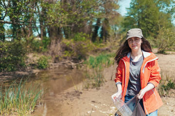 A young female volunteer collects garbage and plastic bottles near the river in a garbage bag. The concept of Earth Day and ecology and environmental protection