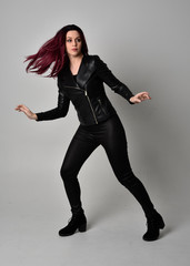 Full length portrait of  girl with red hair wearing black leather jacket, pants and boots. Standing pose, isolated against a grey studio background.