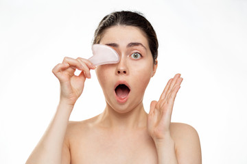 Gua Sha Massage. A young woman holds a quartz gua Sha stone in her hand, covering her eyes and opening her mouth in surprise. White background. Concept of traditional medicine and cosmetology