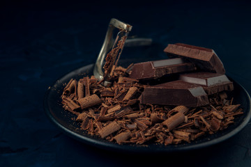 Pieces of chocolate, chocolate shavings and peeler in black plate on dark textured background.