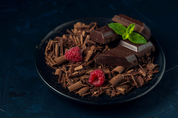 Pieces of chocolate, chocolate shavings, raspberry and mint in black plate on dark textured...