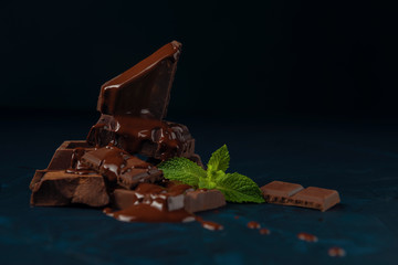 Chocolate pieces and mint leaves poured with liquid chocolate on dark blue textured background.